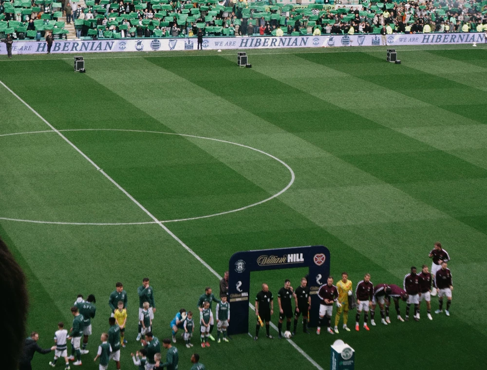 Die Mannschaften laufen im Stadion auf den Rasen ein, während auf der Tribüne im Hintergrund grüner Rauch und Flammensäulen für eine spektakuläre Kulisse sorgen.